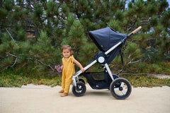 Picture of toddler baby holding purple flowers standing in front of Bumbleride Era reversible stroller in Dusk on dirt trail. New Collection 2022 - Global