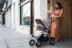 Picture of mother holding child to chest while standing in front of wood door with Bumbleride Era reversible stroller in Sand in front of them. New Collection 2022. Global